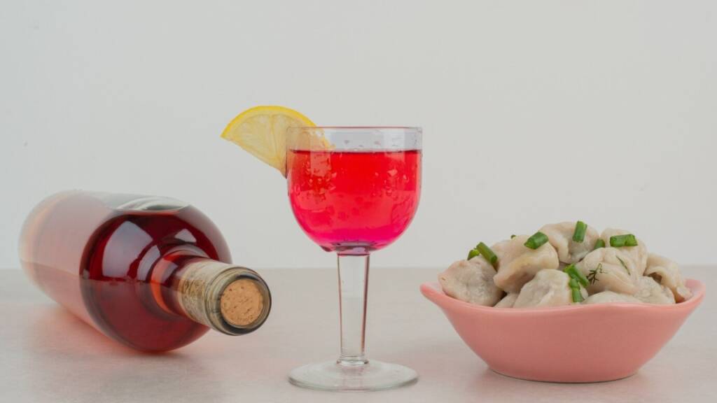 Red cocktail with lemon garnish, dumplings, and a bottle of brandy